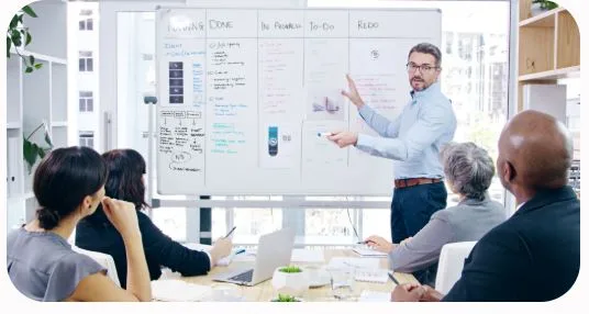 A professional male Business Analyst or facilitator standing and pointing to a large whiteboard containing multi-column process maps, user story criteria, and UI wireframe diagrams, while presenting to an attentive corporate team seated in a brightly lit meeting room.